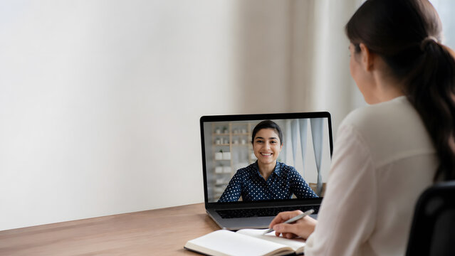 Woman having video call meeting on laptop with smiling colleague on screen while taking note at home