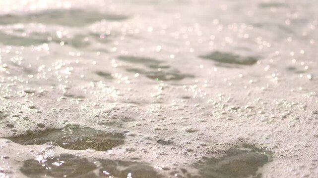 Close-up macro view of shimmering sea foam and tiny glistening bubbles washing over wet sand under bright daylight, capturing a tranquil mood.