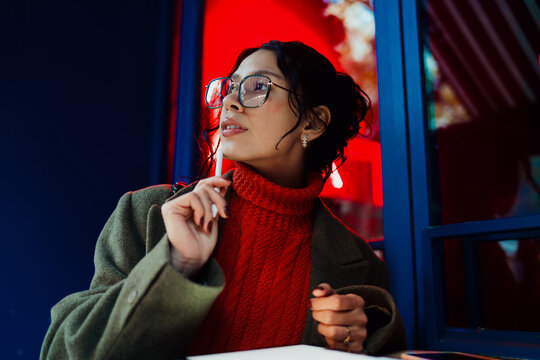 Pensive mixed race female creative thinking about new project while holding stylus, remote work inspiration, brainstorming idea, freelancer, digital nomad, startup culture in city night, glasses.