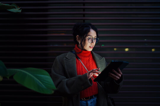Focused multiracial woman in glasses and coat using digital tablet outdoors at night, browsing internet and working on startup, tech savvy professional, mobile office, dark background.