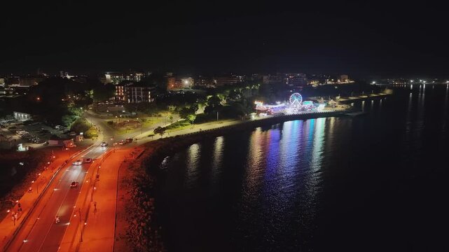 Scenic nighttime 4K drone video shot from near the iconic wooden windmill along the Nesebar isthmus road leading towards the illuminated city. Showcases cars driving on the street, coastal promenade, 