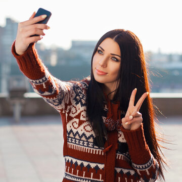 Attractive young brunette woman in a patterned knitted sweater taking a selfie with her smartphone while showing a victory peace sign outdoors in the city on a bright sunny day.