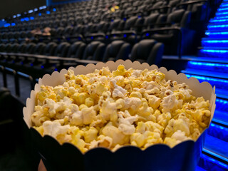 A bucket of popcorn sits on a cinema seat armrest, facing a blank movie screen, with rows of empty theater seats nearby © Татьяна Швец