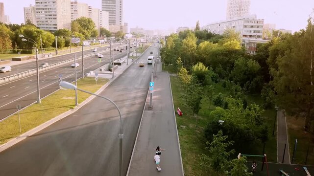 Aerial drone view of young couple riding longboards on urban path along city highway with green trees on warm sunny summer day