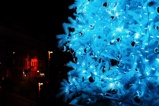 Bright blue and white Christmas tree illuminated at night with colorful lights in the background
