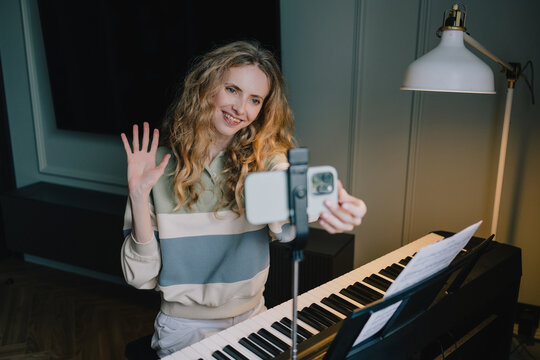 Smiling woman waving at a smartphone beside a digital piano with sheet music at home, connecting online for remote music learning and piano education