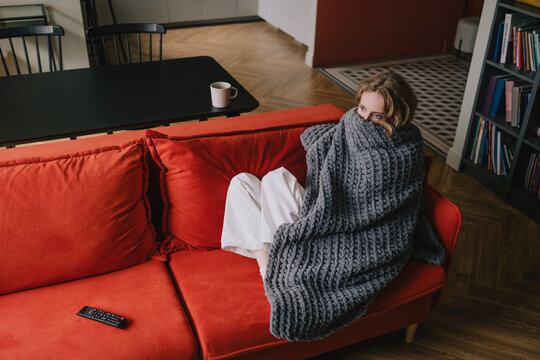 Woman wrapped in a thick blanket to keep warm while sitting on a sofa at home