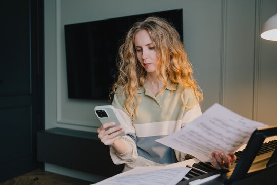 Woman using a smartphone while studying sheet music at a digital piano at home, combining mobile technology with independent music learning and practice