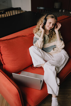 Smiling woman wearing headphones with a laptop while relaxing on a sofa at home, enjoying digital content, online learning and audio entertainment