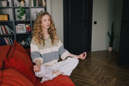 Woman meditating cross-legged on a sofa with closed eyes at home, practicing mindfulness, relaxation and self-care for calm and emotional wellbeing