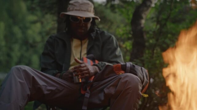 Young adult African American man sitting in folding chair by campfire, holding metal mug and talking, camera tilting up. Video suitable for outdoor recreation or camping gear promotions
