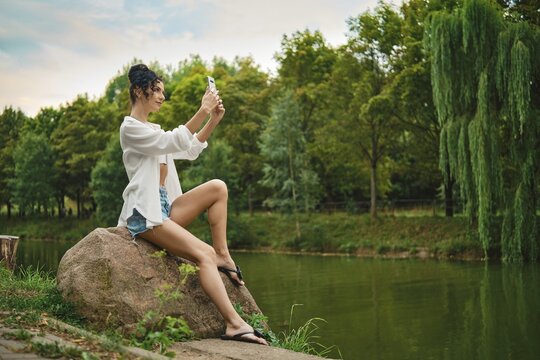 Woman sits on rock next to river in park. She smiles while taking selfie with her phone. Trees surround the area with green leaves. Sky is partly cloudy. She wears shorts and a shirt