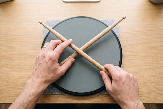 Hands holding wooden drumsticks crossed over a grey circular drum practice pad