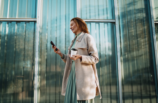 Successful woman scrolling smartphone holding coffee cup standing by glass office building. Mature female professional checking mobile application notifications during morning break. Urban scene.