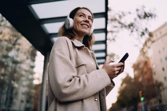 Smiling Caucasian woman enjoying music in headphones at bus stop, happy traveler using smartphone app, urban city lifestyle and digital connectivity, authentic representation, real people.