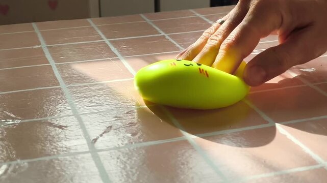 Hand gently presses a bright yellow squishy toy resembling a dumpling on a glossy tiled surface, showcasing playful interaction and tactile enjoyment in a well-lit indoor setting