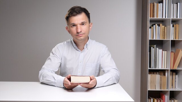 young adult man holding a book while sitting at a white table with a bookshelf in the background, representing education, learning, and academic research
