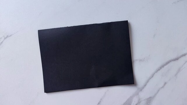 Two childs hands hold a black sheet of paper over a light marble surface, paper craft, folding preparation and creative handmade activity.