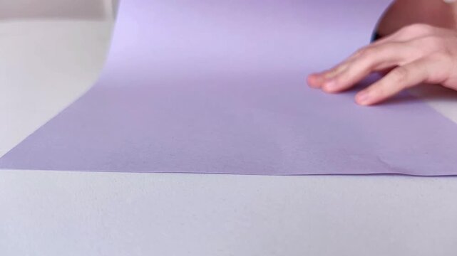 Two childs hands fold a purple sheet of paper on a light marble surface, paper craft, creative activity and handmade design.