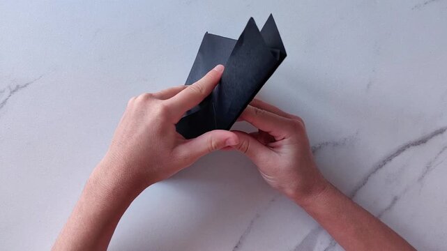 Two hands hold and fold a black sheet of paper into a geometric form, paper craft, origami and handmade creative activity.