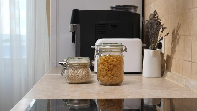 Person's hand is placing a glass jar filled with quinoa on a modern kitchen counter next to another jar of pasta, with a coffee machine and toaster in the background