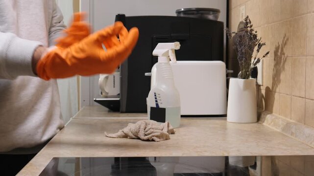 Person is putting on bright orange protective gloves, getting ready to clean and disinfect the kitchen countertop with a spray bottle and a cloth, focusing on hygiene and household tasks