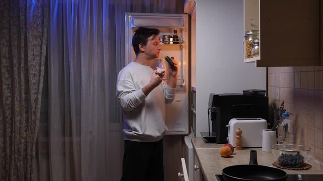 Young man standing by an open refrigerator in a dark kitchen, greedily eating a large sandwich and holding a cucumber, indulging in an unhealthy late night snack