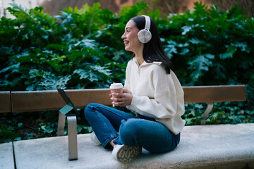 Obraz premium Smiling woman in headphones looking away while using tablet on park bench with cup of coffee. Cheerful girl student or freelancer enjoying work outdoors, remote job and digital connectivity.