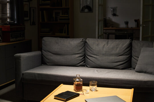 Alcohol decanter and glass sitting on coffee table in front of empty sofa in dark room, representing loneliness or substance abuse. Image suitable for mental health awareness campaigns
