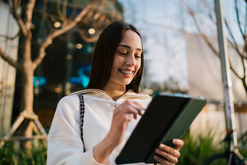 Close up of happy young woman with braces using digital tablet with stylus outdoors. Girl student or creative freelancer working remotely, connectivity and modern technology lifestyle concept. © BullRun