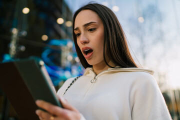 Surprised young woman with dental braces looking at digital tablet in urban city setting. Excited girl winning or receiving Great news on mobile device outdoors, achievement and success concept. © BullRun