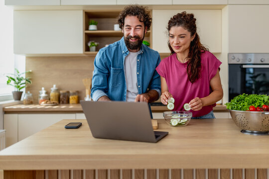 Young couple cooking together in bright kitchen while following recipe on laptop