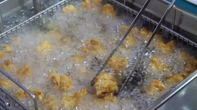 Close-up stock footage of crispy, golden-brown chicken pieces or fritters being deep-fried in hot, bubbling oil inside a commercial metal fryer basket. Metal tongs are used to stir and flip the food t