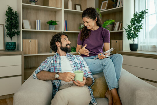 Couple relaxing at home on sofa sharing a happy conversation while holding coffee and notebooks