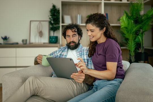 Couple relaxing on couch browsing tablet together at home with coffee and smiles