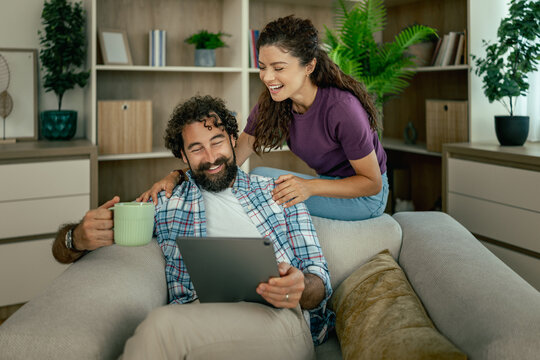 Couple relaxing on sofa at home enjoying tablet and coffee together, smiling and cozy