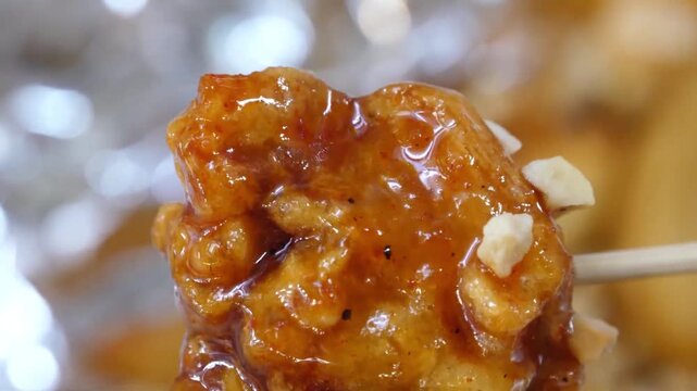 Macro shot of a boneless bite-sized piece of Korean fried chicken (Dakgangjeong) held on a wooden toothpick. The crispy chicken is heavily glazed in a glossy, sticky sweet and spicy gochujang sauce an