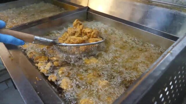 close-up shot of a chef wearing blue gloves using a mesh skimmer to lift freshly fried, crispy golden-brown chicken nuggets from a bubbling commercial deep fryer filled with hot oil. The video capture