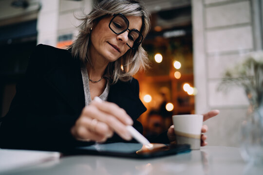 Close up of mature woman using stylus pen to sign digital document on tablet at outdoor cafe, focus on hand interaction with mobile screen, secure online transaction and banking concept