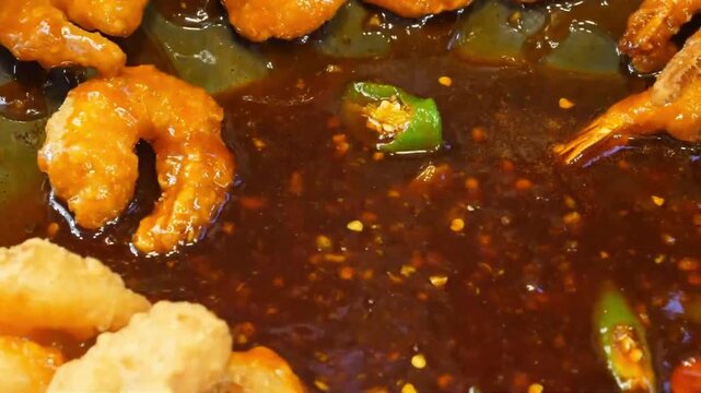 Close-up stock video of battered orange shrimp frying and simmering in a bubbling, deep brown savory chili sauce. The dish is being prepared in a large wok or pan, featuring visible red chili flakes a