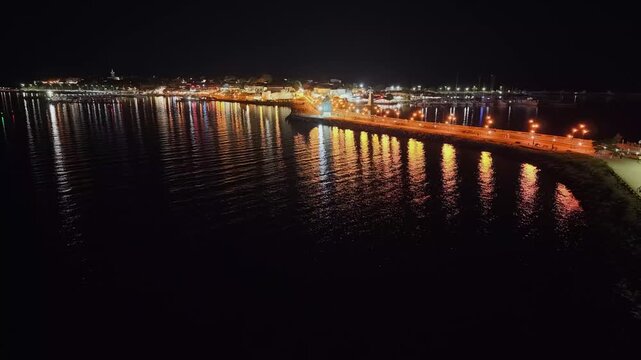 Nighttime aerial 4K drone video of the illuminated isthmus in Nesebar, Bulgaria. Features golden streetlight reflections shimmering on the calm Black Sea, with the old town cityscape and coastal prome