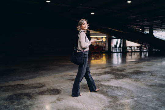 Smiling mature Caucasian woman walking and holding smartphone in empty hall, female looking back at camera while using mobile app, authentic urban portrait with copy space in dark interior.