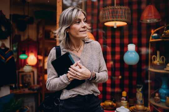 Side view of mature woman with tablet looking at shop window display in city, curious female freelancer browsing urban environment, modern digital lifestyle and remote work concept.