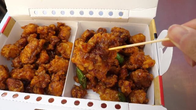 close-up shot of a person using a wooden toothpick to pick up a piece of crispy, glazed Korean fried chicken (Dakgangjeong) from a white cardboard takeout delivery box. The box is divided into two sec