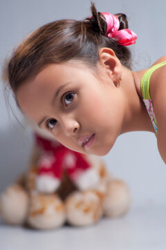 Curious young girl with pink hair ribbons leaning forward and looking closely with teddy bear blurred in background