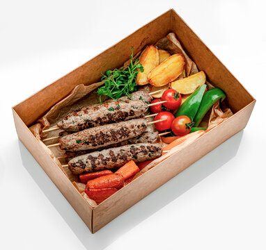 Grilled meat skewers with potatoes and vegetables in box. Grilled minced meat skewers are arranged with roasted potatoes, carrots, bell peppers, and tomatoes in a brown paper box.