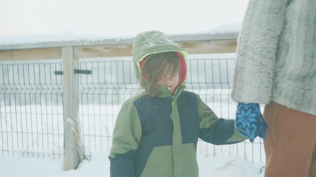 Toddler in a green hooded jacket smiling while holding his mothers hand on a snowy walkway