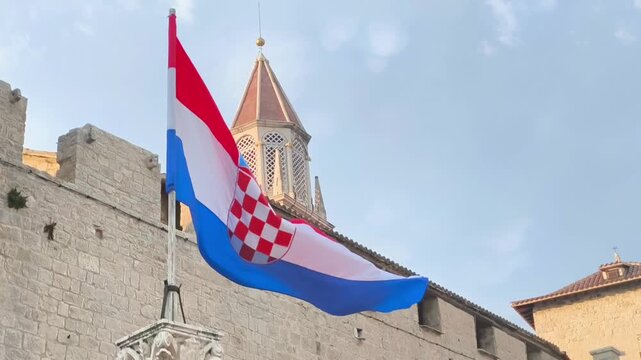Croatian flag waving in the breeze against a historic stone building with a tower and cloudy sky, showcasing vibrant colors and intricate design elements