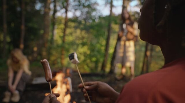 African American young adult man roasting marshmallow and eating grilled sausage by campfire with multiethnic friends in forest. Ideal for outdoor recreation and camping gear promotions