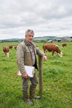 Male farmer in gray jacket stands beside wooden post holding clipboard, with brown and white cows grazing in green pasture and farm buildings in the background